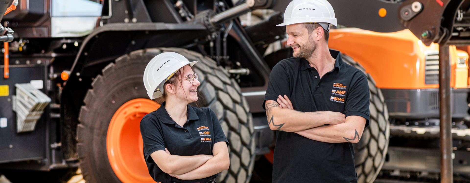 Zwei Bauarbeiter in schwarzen Uniformen und weißen Schutzhelmen stehen sich lächelnd und mit verschränkten Armen vor einer großen Industriemaschine mit schwarzen Reifen gegenüber.
